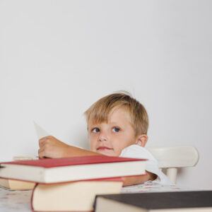 Menino pequeno sentado à mesa com livros, folheando páginas com expressão pensativa, ilustrando dificuldades de leitura e aprendizado